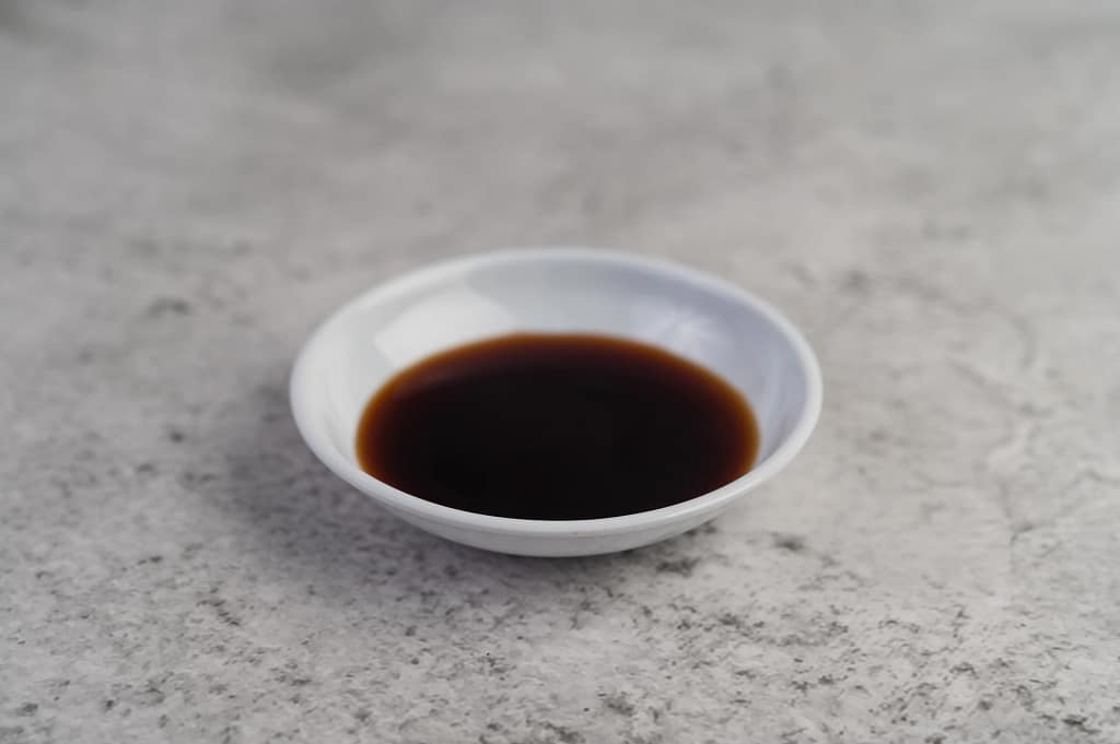 black-sauce-small-white-cup-placed-cement-floor_11zon Teriyaki sauce served in a small white dish on a textured surface.