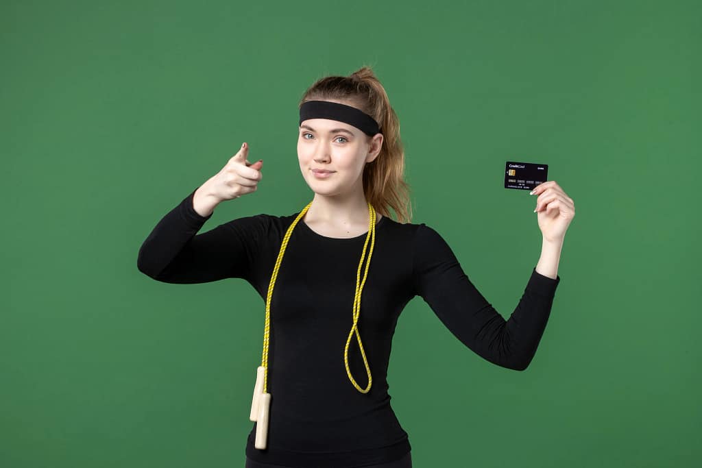 Young woman in fitness clothing holding a credit card while posing confidently against a green background.