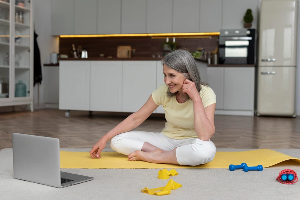 A woman exercising at home on a yoga mat while learning from her online personal trainer for seniors on her laptop.