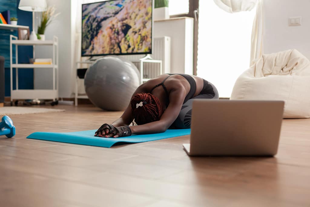 Online General Fitness Trainers A woman stretching on a yoga mat at home while following an online session with general fitness trainers on a laptop.