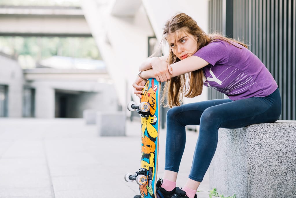 A teenage girl is taking a break from skateboarding, leaning on her colorful skateboard.
