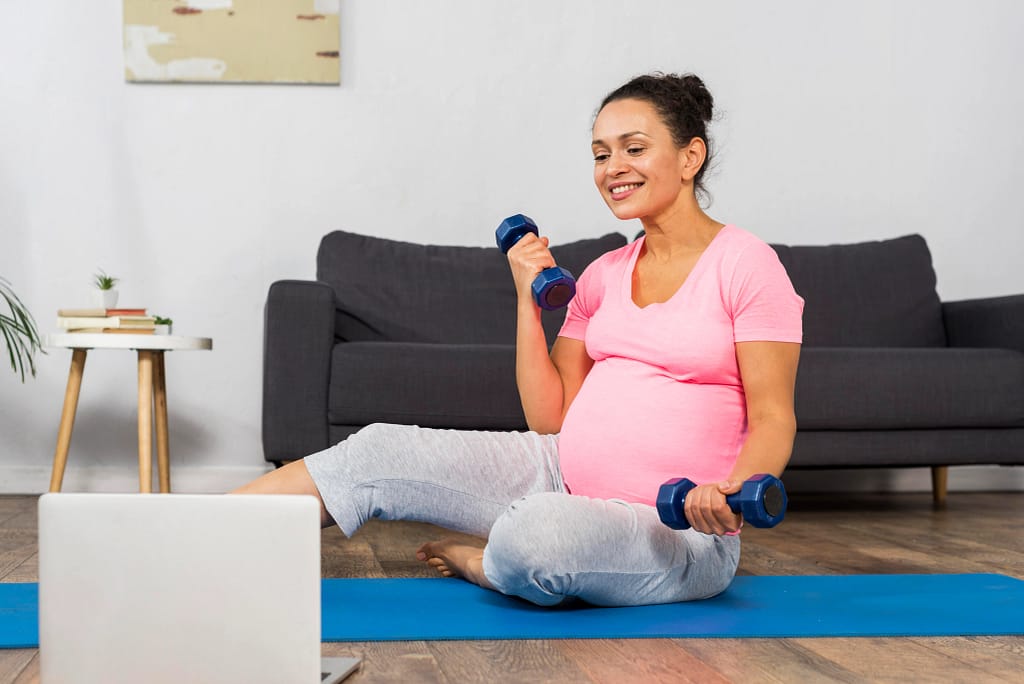 Pregnant woman exercising at home with dumbbells while following guidance from online personal trainers for women.